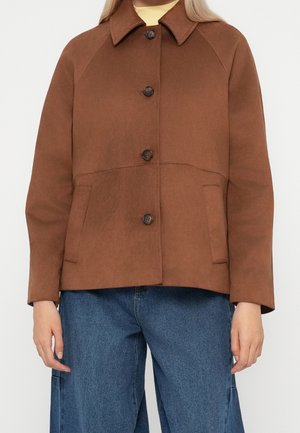 Kobieta w brązowej kurtce zapinanej na guziki z bocznymi kieszeniami i niebieskich dżinsach, stojąca na tle jasnego, jednolitego tła.