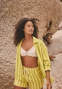 Camicia a righe gialla chiara con bottoni, bralette bianca testurizzata, pantaloni corti a righe coordinati con coulisse, sfondo roccioso naturale.