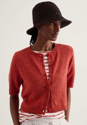 Donna che indossa un cardigan rosso a maniche corte con bottoni sopra una camicia a righe rosse e bianche, con un cappello a secchiello lavorato all'uncinetto di colore marrone scuro.