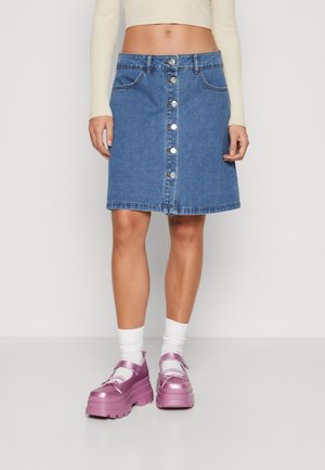 Mujer con falda vaquera azul con botones delanteros, calcetines blancos y zapatos Mary Jane de plataforma gruesa rosa, de pie contra un fondo liso.
