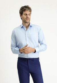 Camicia azzurro chiaro a maniche lunghe con colletto a punta, taschino singolo sul petto e un branding discreto, abbinata a pantaloni blu navy.