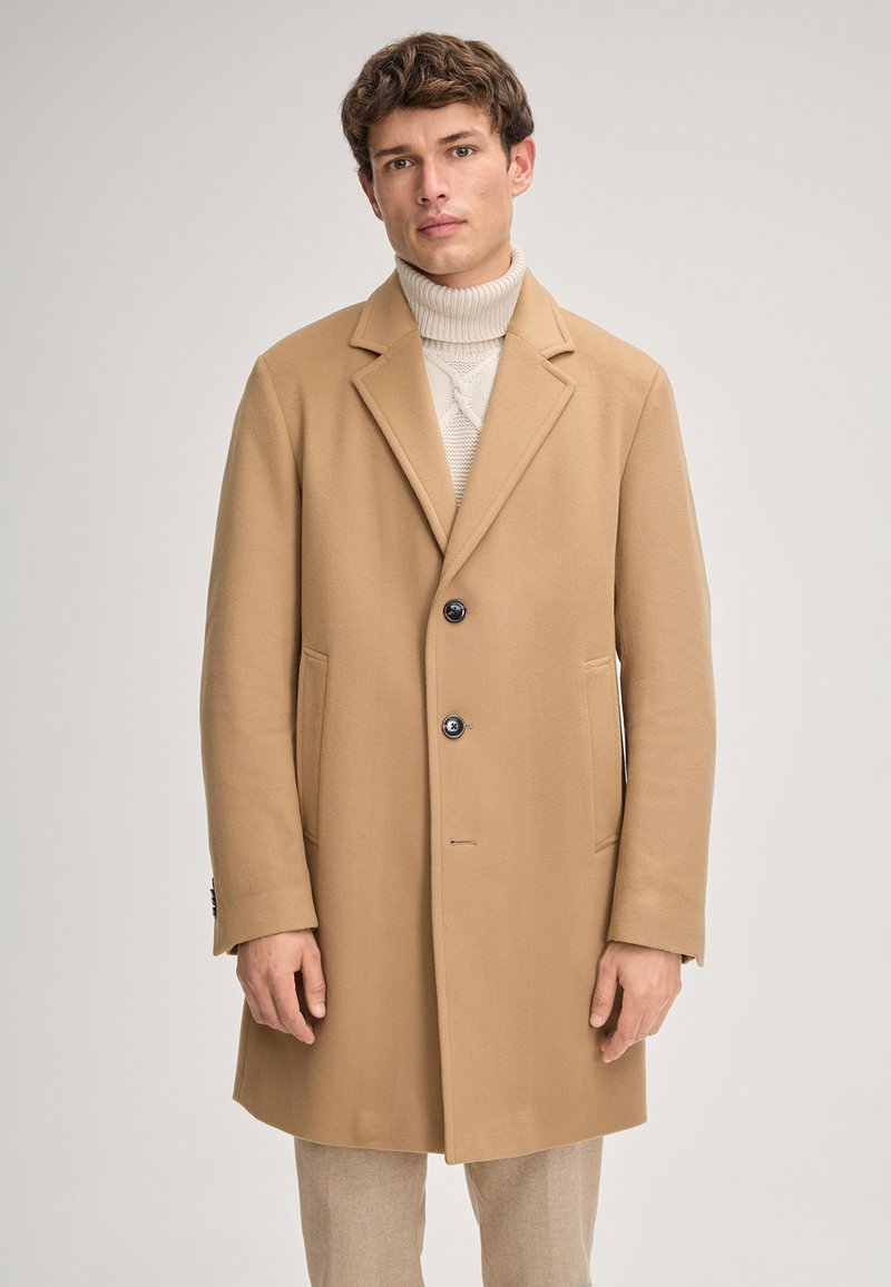 Jonge man in een beige overjas over een crèmekleurige coltrui en lichtgekleurde broek tegen een effen achtergrond.