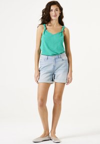 Top verde claro de tirantes con detalles de nudos, combinado con shorts de mezclilla azul claro con dobladillos enrollados. La modelo lleva unos zapatos planos plateados.