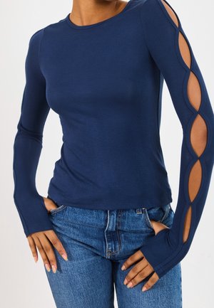 Žena v námořnicky modrém topu s dlouhými rukávy a otevřenými oválnými výřezy na jednom rukávu, v kombinaci s modrými džínami. Ruce má založené v kapsách.