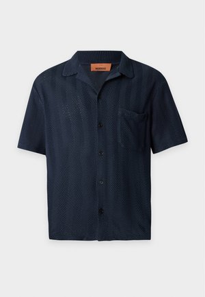 Mann steht und trägt ein schwarzes kurzärmeliges Hemd mit Knöpfen, hellblaue Jeans und weiße Turnschuhe vor einem schlichten weißen Hintergrund.