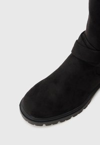 Schwarze Wildleder-Stiefelette mit runder Zehenpartie, gewickeltem Riemen-Detail und einer strukturierten Gummisohle mit Rutschmustern.