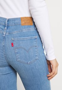 Helle blaue Jeans mit hoher Taille, featuring ein Lederetikett auf der Rückseite und einem roten Tab an der rechten Tasche, hervorgehobene Nähte.