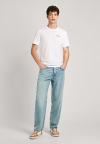Camiseta blanca de algodón con un pequeño logo negro, combinada con unos jeans de denim azul claro y zapatillas marrones y blancas. Ajuste y diseño casual.