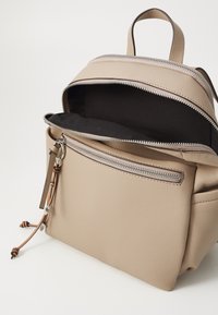 Malý béžový kožený batoh s otevřenou hlavní přihrádkou, přední kapsou na zip a dvěma koženými třásněmi s kovovými nýty na zipovém táhle.