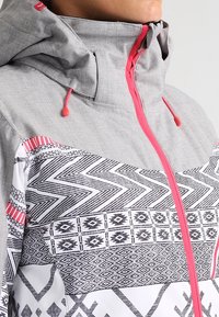 Graue Jacke mit gemusterter Vorderseite, die schwarze geometrische Designs und rosa Akzente aufweist. Sie hat eine Kapuze und einen Reißverschluss mit Kordelzügen.