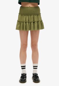 Olijfgroene gelaagde rok van zachte stof, met een smocktail en een gerimpelde zoom. Gekozen met zwarte schoenen en gestreepte sokken.