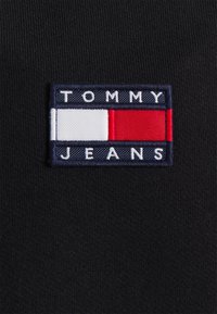 Tejido negro que presenta un parche rectangular con el logo de Tommy Jeans. El parche muestra el nombre de la marca en blanco sobre fondo azul marino, con rayas horizontales en rojo y blanco.