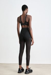 Top desportivo preto e leggings de cintura alta feitas de material suave; apresenta painéis de malha. O calçado é composto por sapatilhas pretas de desporto com solas texturizadas.