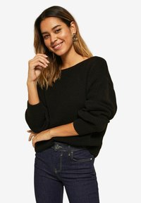 Pull en tricot noir avec un large col et des manches retroussées, associé à un jean en denim foncé avec des surpiqûres jaunes et une poche avant.