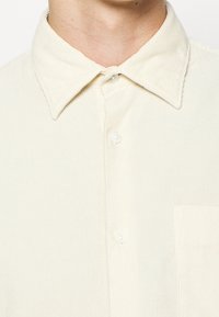 Camicia in corduroy color crema con colletto a punta, tre bottoni e taschino sul petto. Tessuto a coste con linee verticali.