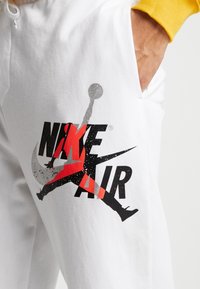 Bílé teplákové kalhoty s velkým černo-červeným nápisem "NIKE AIR" se siluetou skákající postavy a šedým logem Swoosh.