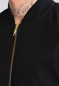 Veste bomber noire avec fermeture éclair à l'avant, dotée d'une fermeture éclair dorée, d'un col côtelé et d'une texture lisse.