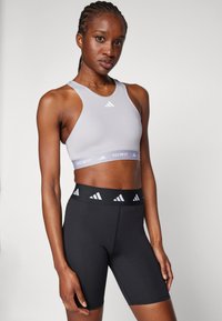 Kvinne med flettehår iført en lys grå Adidas Techfit sports-bh og svarte Adidas høytaljede treningsshorts, stående mot en ensfarget bakgrunn.