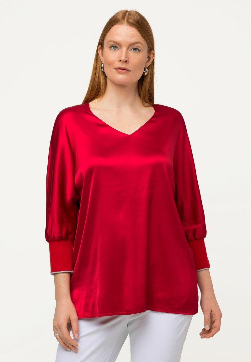 Haut en satin rouge avec un col en V et des manches bouffantes à trois quart. Texture lisse, coupe légèrement ample, et poignets contrastants. Pantalon blanc.