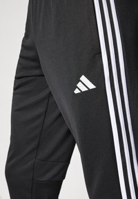 Svarte treningbukser med tre hvite striper langs siden og en hvit trekantet logo. Materialet ser glatt ut med en teksturert lomme.