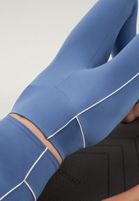 Visoko pasovi modri legingi iz gladkega materiala z belimi obrobami ob šivih, razstavljeni na strukturirani črni jogijski podlagi.