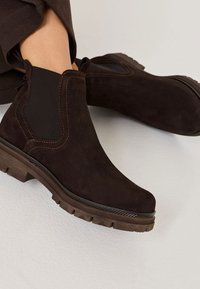 Brązowe zamszowe botki za kostkę z elastycznymi panelami po bokach, grubymi gumowymi podeszwami i kontrastującymi przeszyciami. Okrągły czubek i teksturowane wykończenie.