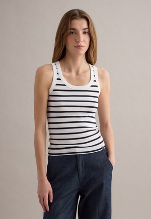Junge Frau trägt ein weißes Tanktop mit schwarzen horizontalen Streifen und dunkelblauer Hose, steht mit einer Hand in der Tasche vor einfarbigem Hintergrund.