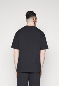 Man with braided hair tied in a bun wearing a plain black short-sleeve shirt and black pants, standing facing away on a white background.