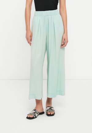 Pantalon ample vert clair plissé avec une taille élastique. Porté avec des sandales à enfiler noires et blanches à motifs, semelle plate.
