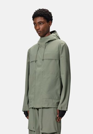 Giacca impermeabile verde chiaro con cappuccio, dotata di due tasche superiori sul petto, orlo regolabile con cordino e dettagli neri.