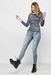 Tamsiai mėlynas ilgomis rankovėmis marškinėlis su gėlių siuvinėjimu, derinamas su šviesiai mėlynomis nusidėvėjusiomis siaurėjimo džinsais ir juodais šnuoruotais auliniais batais.