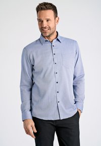 Lichtblauwe overhemden met lange mouwen, kleine borstzak, donkere knopen en een subtiel textuurpatroon. Gecombineerd met zwarte broek.