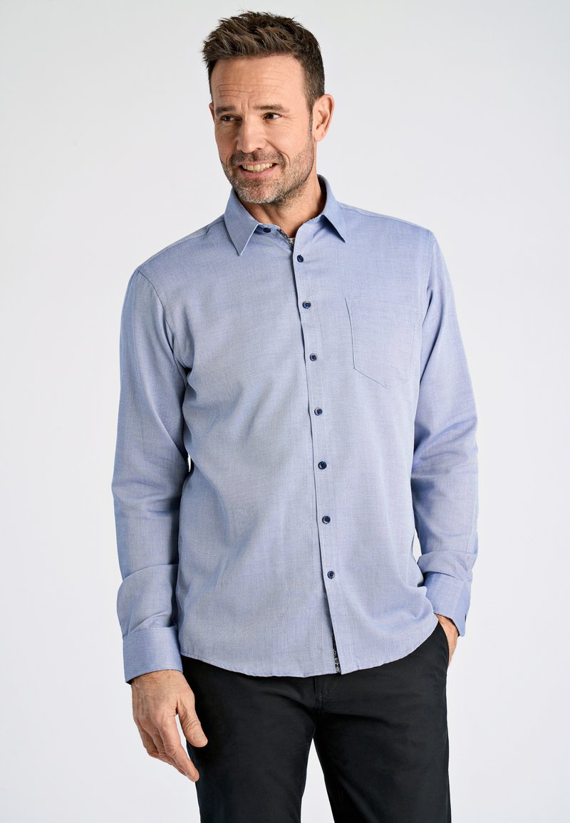 Lichtblauwe overhemden met lange mouwen, kleine borstzak, donkere knopen en een subtiel textuurpatroon. Gecombineerd met zwarte broek.