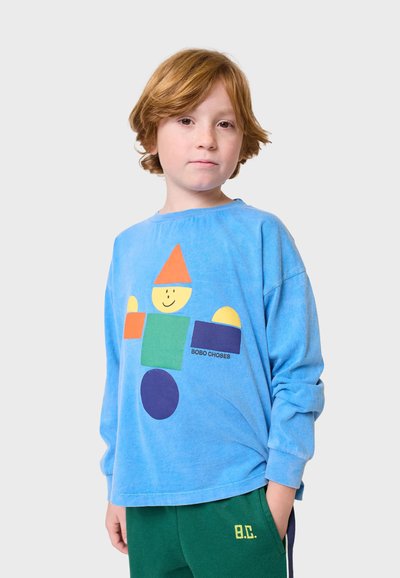 Sininen pitkähihainen paita, jossa on leikkisä geometrinen kuvio, jossa on hymyilevä hahmo keltaisena, vihreänä ja oranssina. Pehmeä kangas, rento istuvuus.