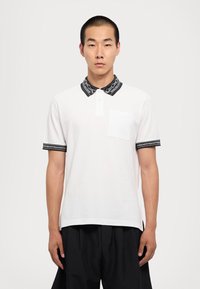 Mladý muž s krátkymi vlasmi, oblečený v bielom polo tričku s čierne vzorovanými golierom a rukávmi, stojaci na pozadí z jednej farby - bielej.