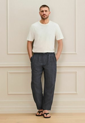 Mann steht vor einer beigen Wand, trägt ein weißes kurzärmeliges Hemd, dunkle Hose mit Kordelzug und schwarze Zehensandalen, die Hände in den Taschen.