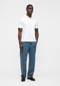 Homme debout, portant un polo blanc à manches courtes, un jean bleu et des chaussures marron foncé, sur un fond blanc uni.