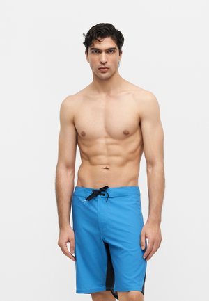 Mišićav muškarac s tamnom kosom u plavim kupaćim gaćicama s crnim vezicama, stoji ispred jednobojne svijetle pozadine.