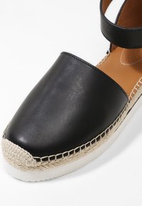 Espadrille de cuero negro con punta redonda, que presenta un borde de yute trenzado y una correa para el tobillo. Textura suave y suela blanca resistente.