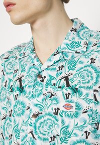 Gros plan d'une personne portant une chemise blanche boutonnée avec des motifs floraux turquoise et des champignons noirs, avec un logo Dickies sur la poche poitrine.
