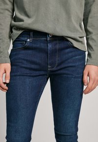 Temno modri daske kavbojke s slim fit krojem, vidnim šivom in sprednjimi žepi. Kombinirane z dolgimi rokavi zeleno srajco.