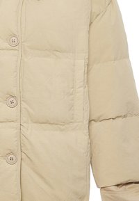 Chaqueta acolchada beige con un acabado texturizado. Presenta un cierre de botón y un bolsillo en el pecho. Suave, cálida y ligera, ideal para climas fríos.