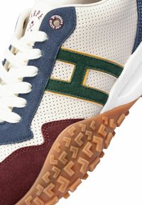 Zapatilla con un upper perforado en blanco, acentos en azul marino y burdeos, logo de ante verde y suela de goma texturizada con un patrón en zigzag.