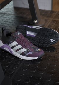 Ζευγάρι μοβ και μαύρα αθλητικά Adidas με λευκές λωρίδες και σχέδιο πεταλούδας, τοποθετημένα σε μαύρο υφασμένο λαστιχένιο δάπεδο.