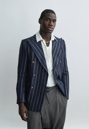 Egy férfi sötétkék csíkos kétsoros blézerben, fehér ingben, szürke nadrágban és ezüst nyaklánccal áll egy egyszerű világos háttér előtt.