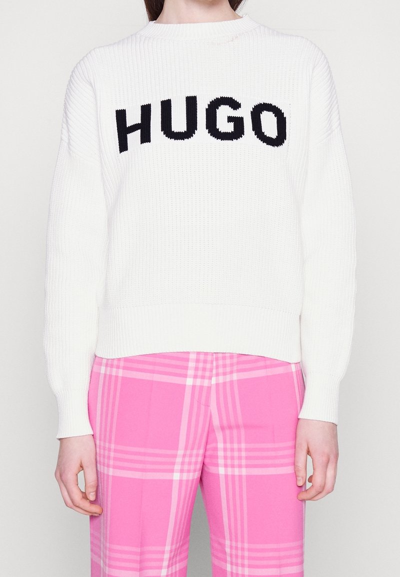 Personne portant un pull en maille blanc avec "HUGO" en lettres noires et un pantalon large à carreaux roses, debout devant un fond uni.