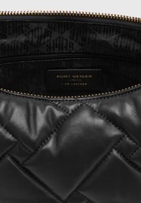 Černá prošívaná kožená taška se zlatým zipem, ukazující vnitřní látkovou podšívku a nálepku s nápisem "Kurt Geiger London I Am Leather."
