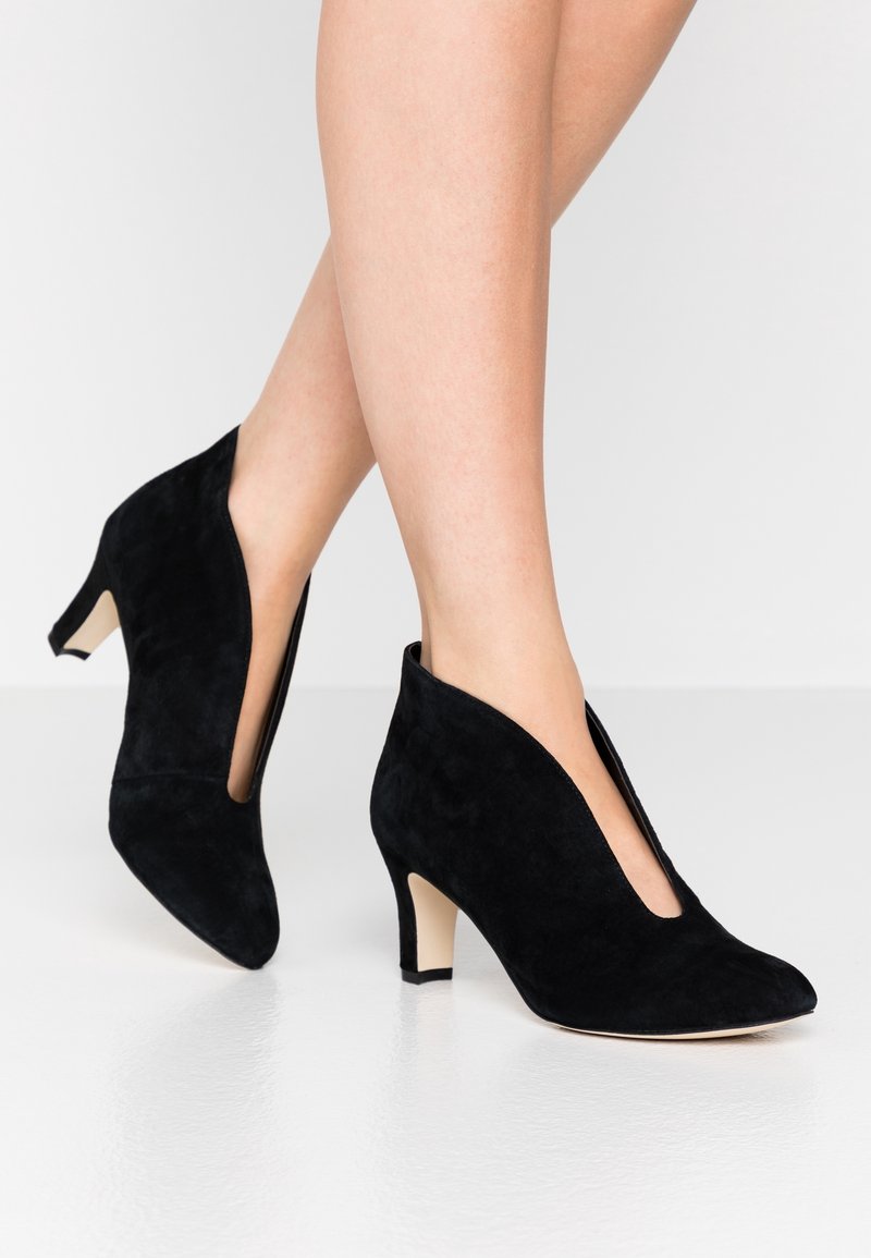 Jambes portant des chaussures à talons hauts en daim noir avec une découpe en V profonde à l'avant, debout sur une surface blanche lisse sur un fond clair uni.
