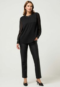 Blusa nera con maniche lunghe trasparenti e scollo decorato, abbinata a pantaloni neri aderenti e tacchi a punta. Texture del tessuto liscia.