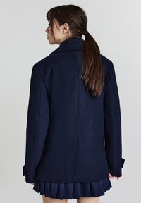 Marineblauer Wollmantel mit Reverskragen, strukturierten Schultern und langen Ärmeln, ausgestattet mit einer glatten Textur und einer taillierten Passform. Faltenrock in Marineblau sichtbar.